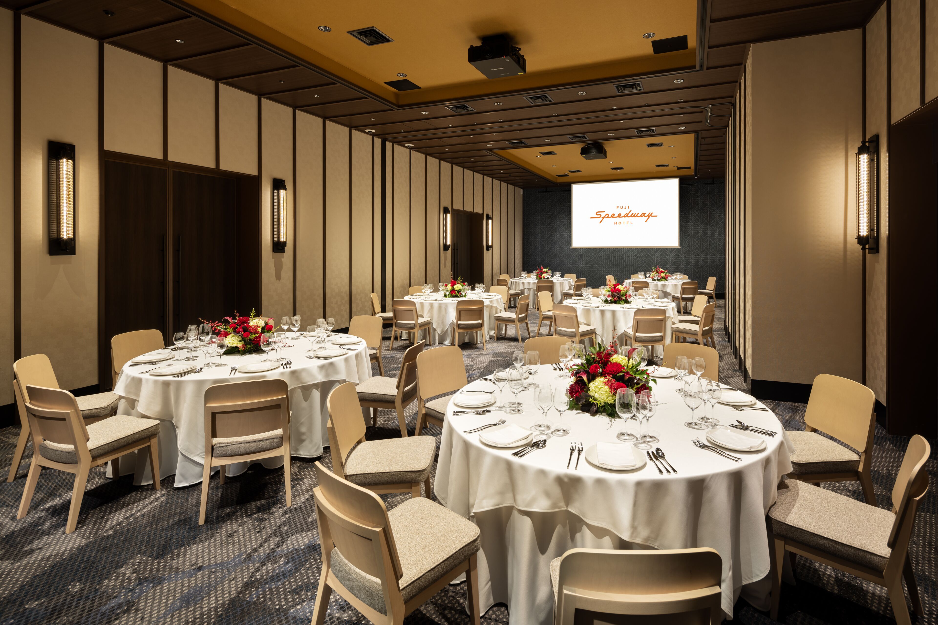 Une salle de conférence dotée de plusieurs tables rondes, chacune recouverte d’une nappe blanche et agrémentée de chaises et de compositions florales.