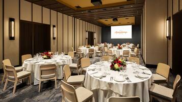 Une salle de conférence dotée de plusieurs tables rondes, chacune recouverte d’une nappe blanche et agrémentée de chaises et de compositions florales.