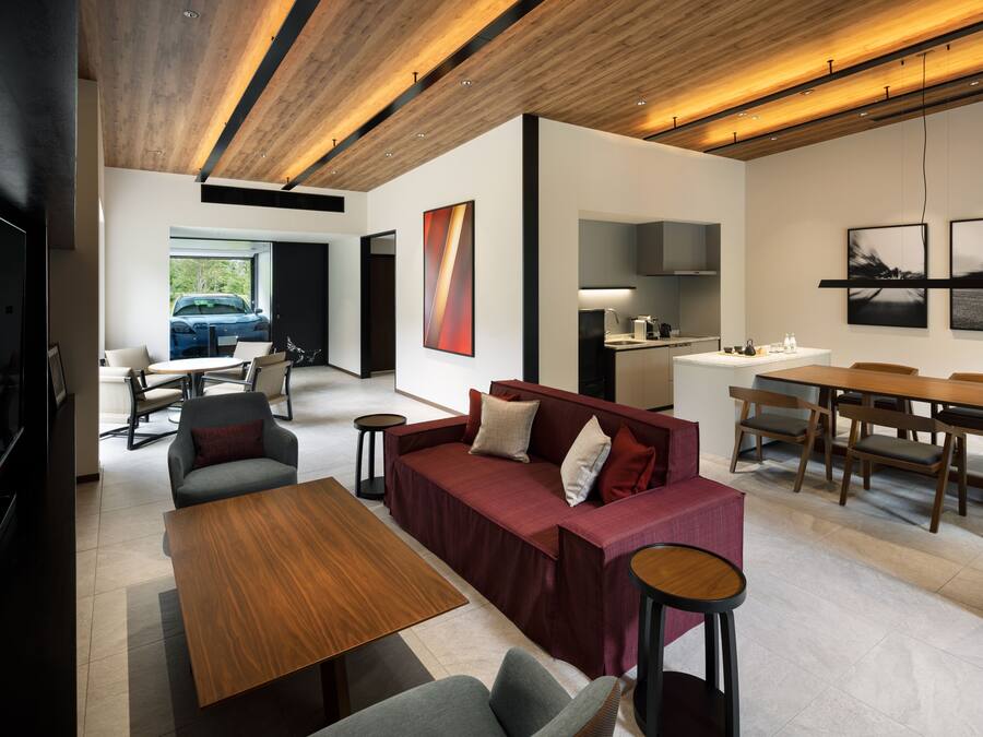 A modern living room with a wooden ceiling, a sofa set, a dining table, and a kitchen area visible through an open door.
