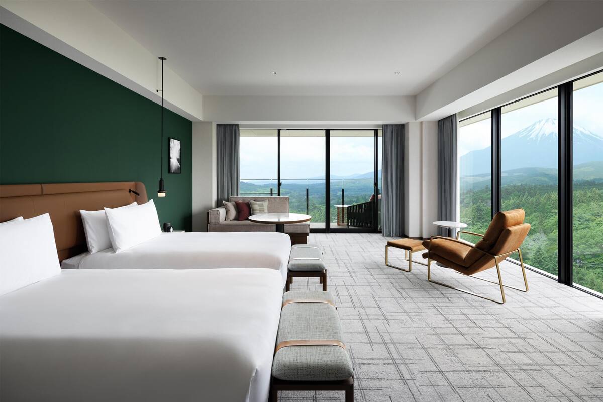 A hotel room with a large bed, a sofa, a small table, and a chair. There is a balcony with a view of a mountain.