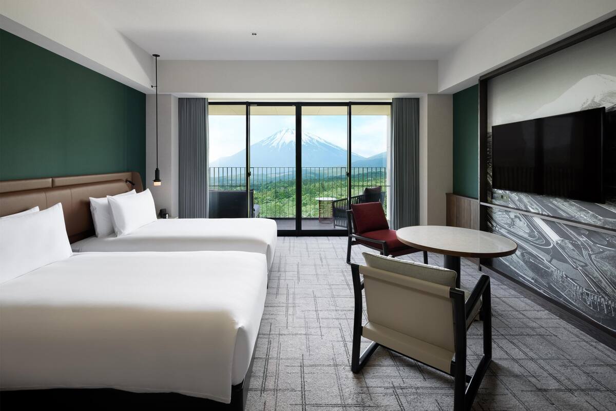 A hotel room with a large bed, a small table, a chair, a TV, and a balcony with a mountain view.
