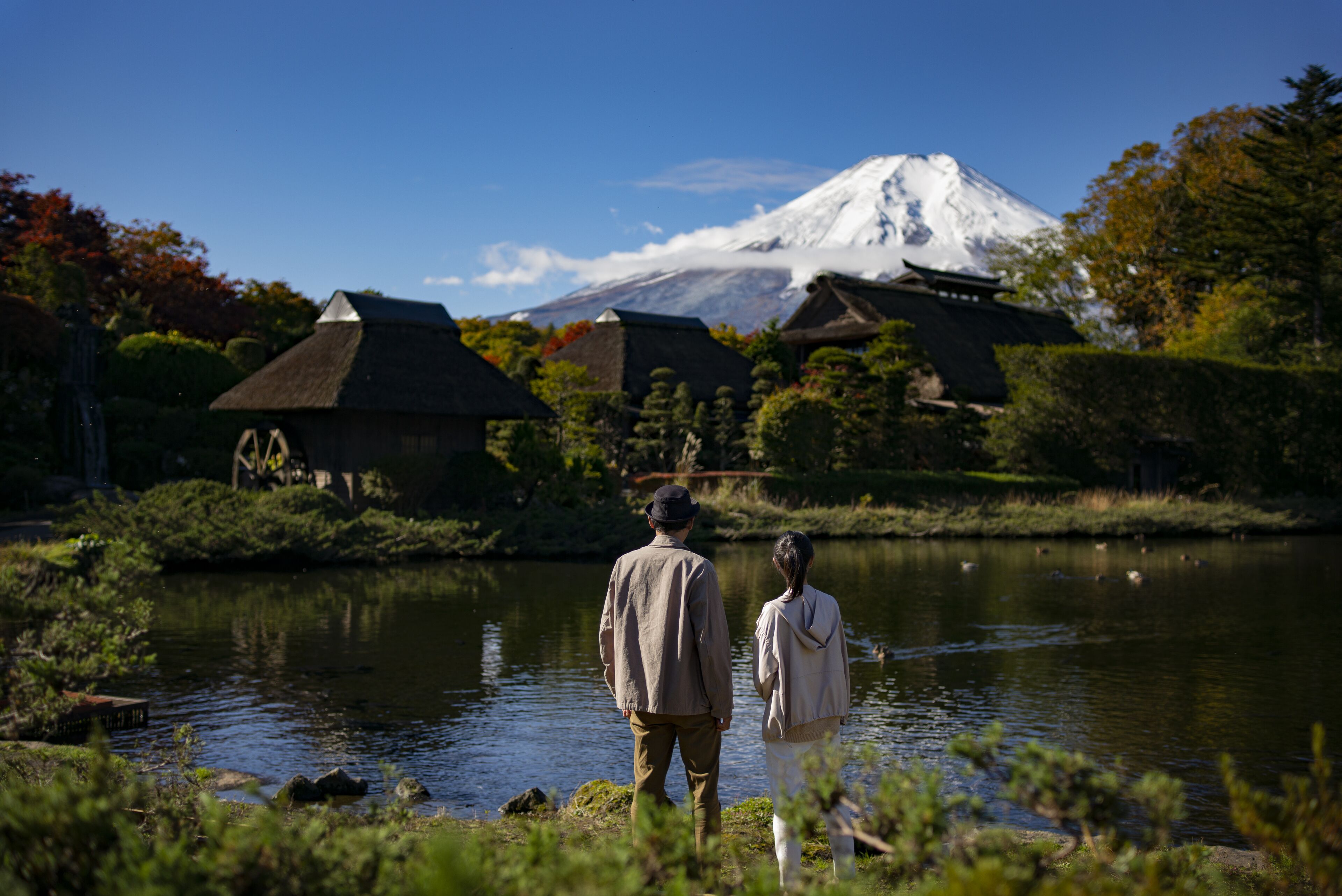 A couple standing by a calm lake with traditional buildings and a snow-capped mountain in the background.