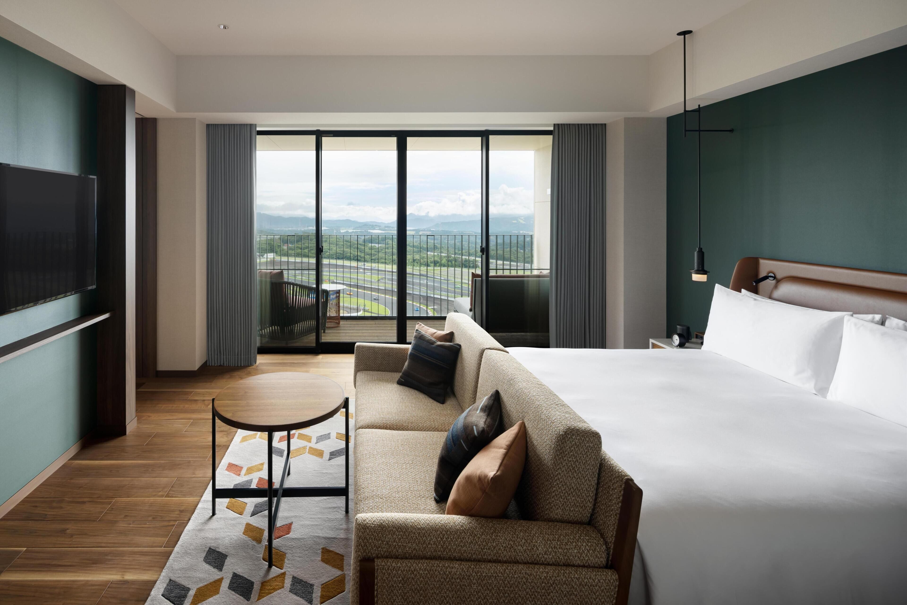 A hotel room with a bed, a sofa, a TV, a balcony with a view, and a dining area.
