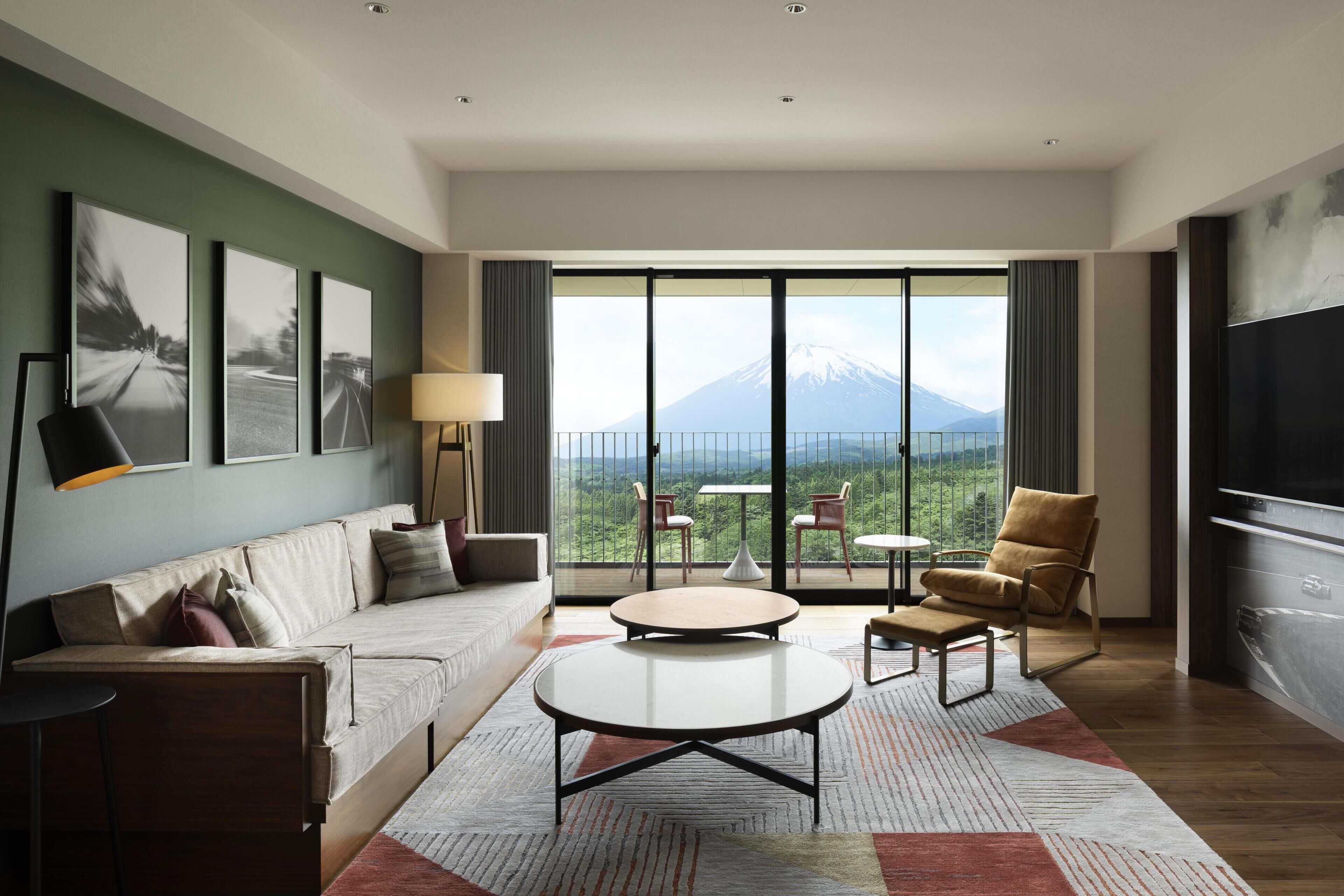 A modern hotel room with a sofa, a round coffee table, a TV, and a balcony with a mountain view.