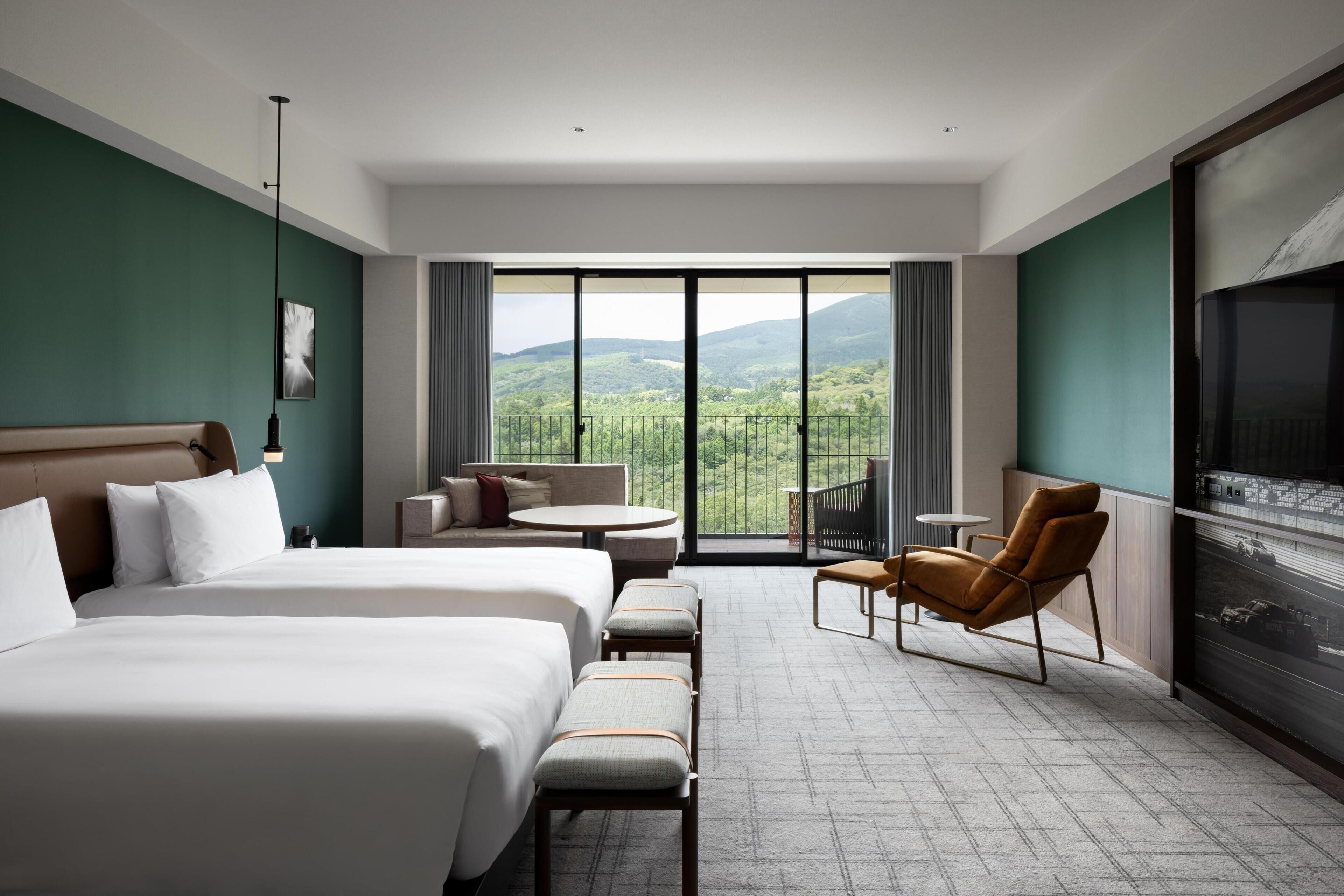 A hotel room with two beds, a sofa, a chair, a TV, and a balcony with a mountain view.