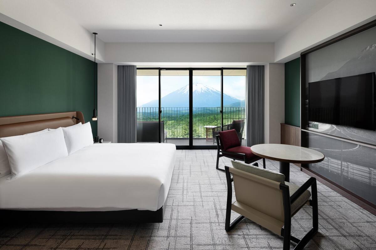 A hotel room with a bed, a chair, a table, a TV, and a balcony with a mountain view.