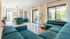 Een ruime zithoek met blauwgroene banken, een vloerlamp en een grote schuifpui naar een balkon.
