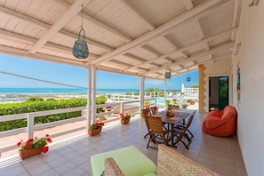 Um alpendre coberto com vista para o mar, uma mesa de jantar, cadeiras e um pufe.