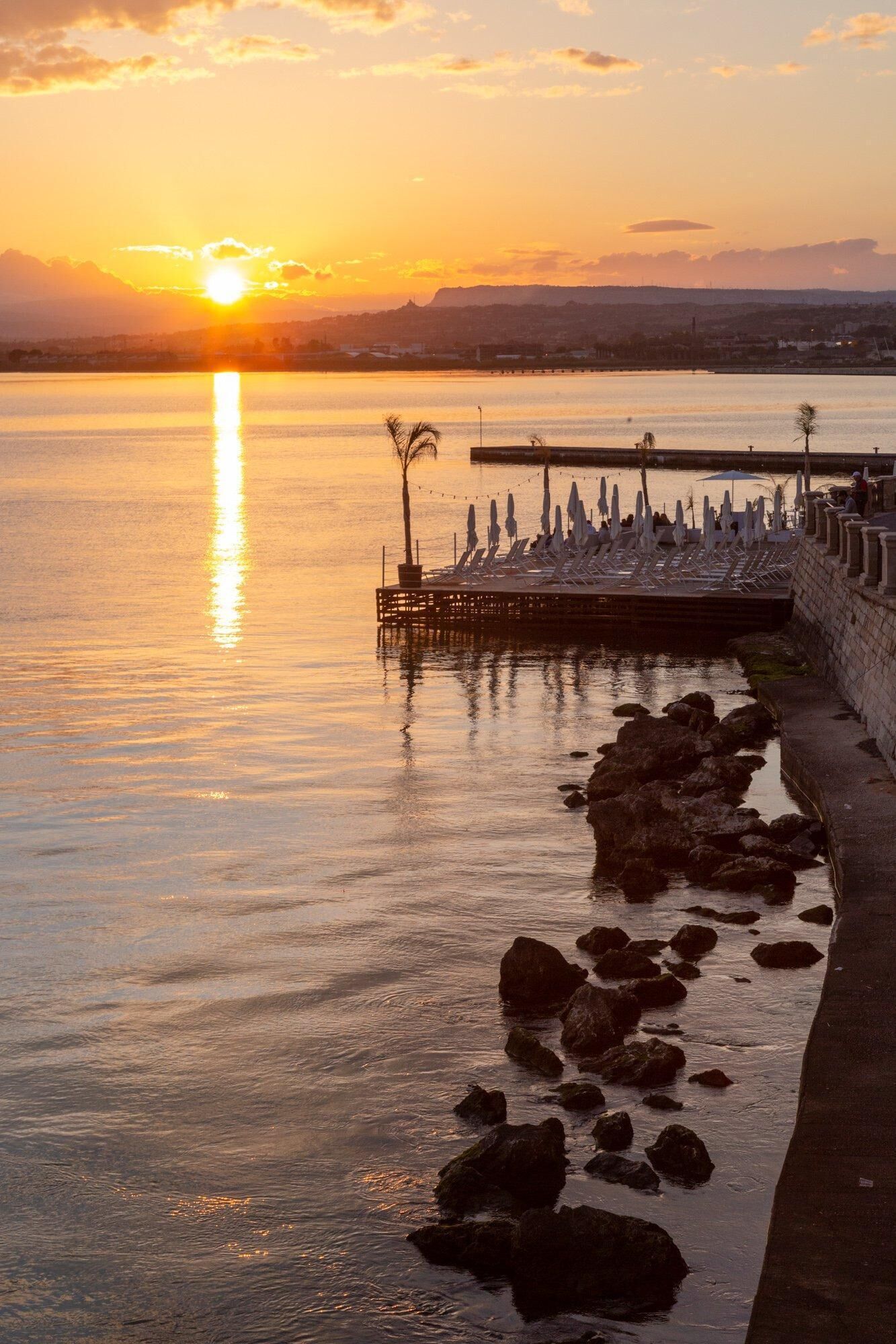 Une scène de bord de mer avec une jetée s’avançant dans des eaux calmes, une rangée de chaises longues et une côte rocheuse.