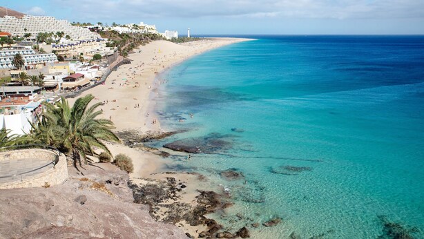 Una playa costera con edificios, agua turquesa cristalina y zonas rocosas.