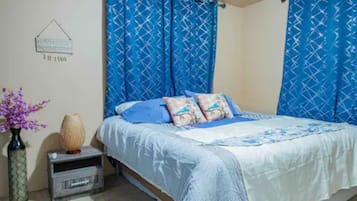 Ein Schlafzimmer mit einem Bett, blauen Vorhängen, einem kleinen Nachttisch mit einer Vase und einer Wanddekoration.