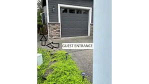 A house with a garage door, a guest entrance sign, and a small garden area with plants and a solar-powered outdoor light. - Home: Sleeps 4, Bedroom, Living, Bath, Office, Patio (Bothell)