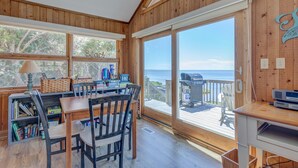 A wooden interior with a dining area, bookshelves, and a view of the ocean through large sliding glass doors. - 7815-Tarhoo, | Soundfront Frisco Retreat | Pool & Sunset Views (Frisco)