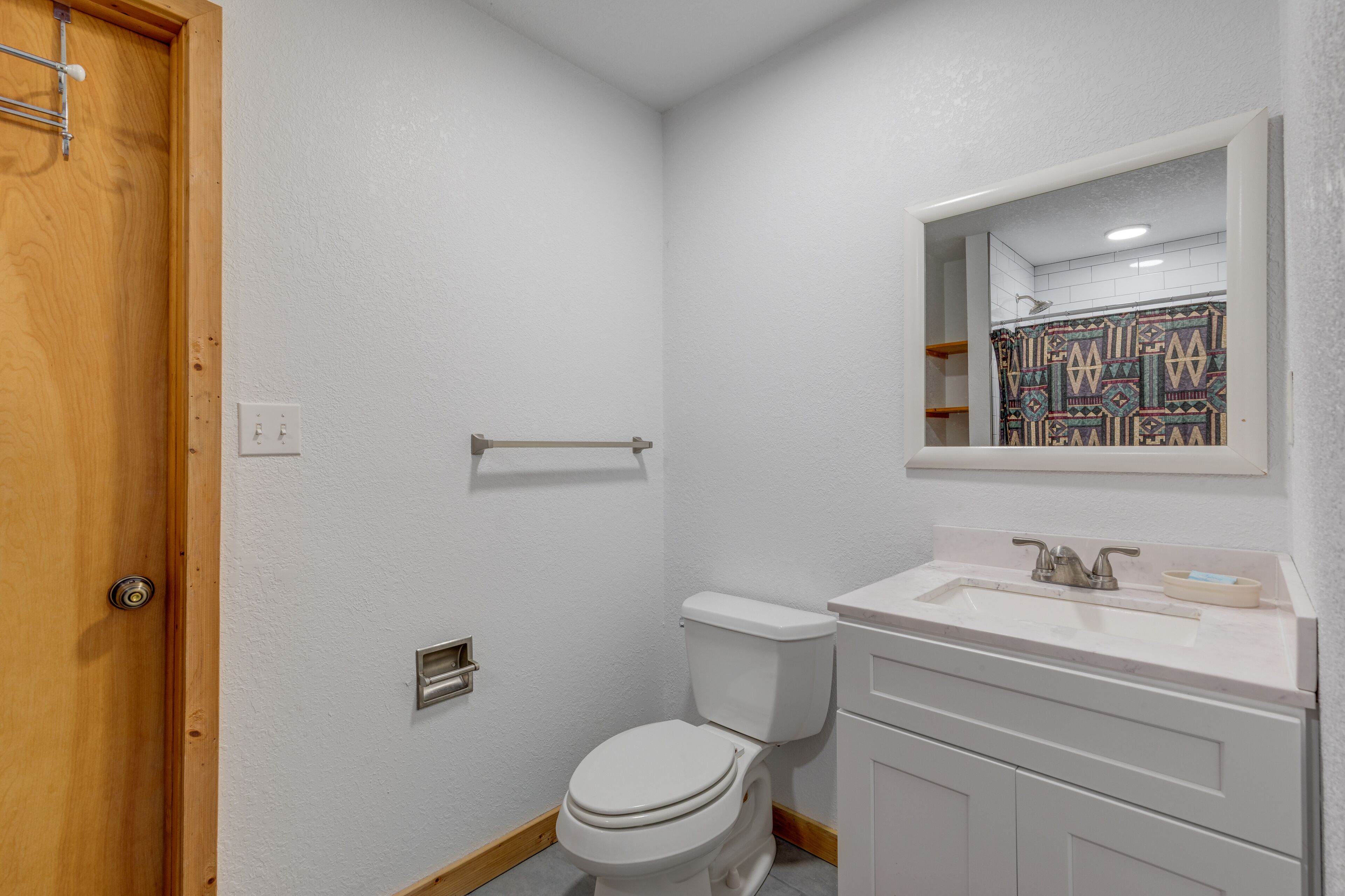 Une petite salle de bain propre, avec des toilettes blanches, un lavabo surmonté d’un miroir et un rideau de douche à motifs géométriques.