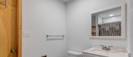 Une petite salle de bain propre, avec des toilettes blanches, un lavabo surmonté d’un miroir et un rideau de douche à motifs géométriques.