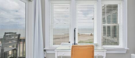 A room with a desk and chair, a window with blinds, and artwork on the wall.