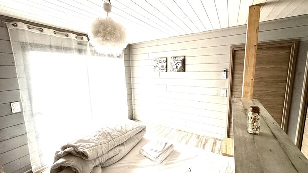 A bedroom with a bed, a window with blinds, a wooden ceiling, and a wall-mounted shelf with a jar.