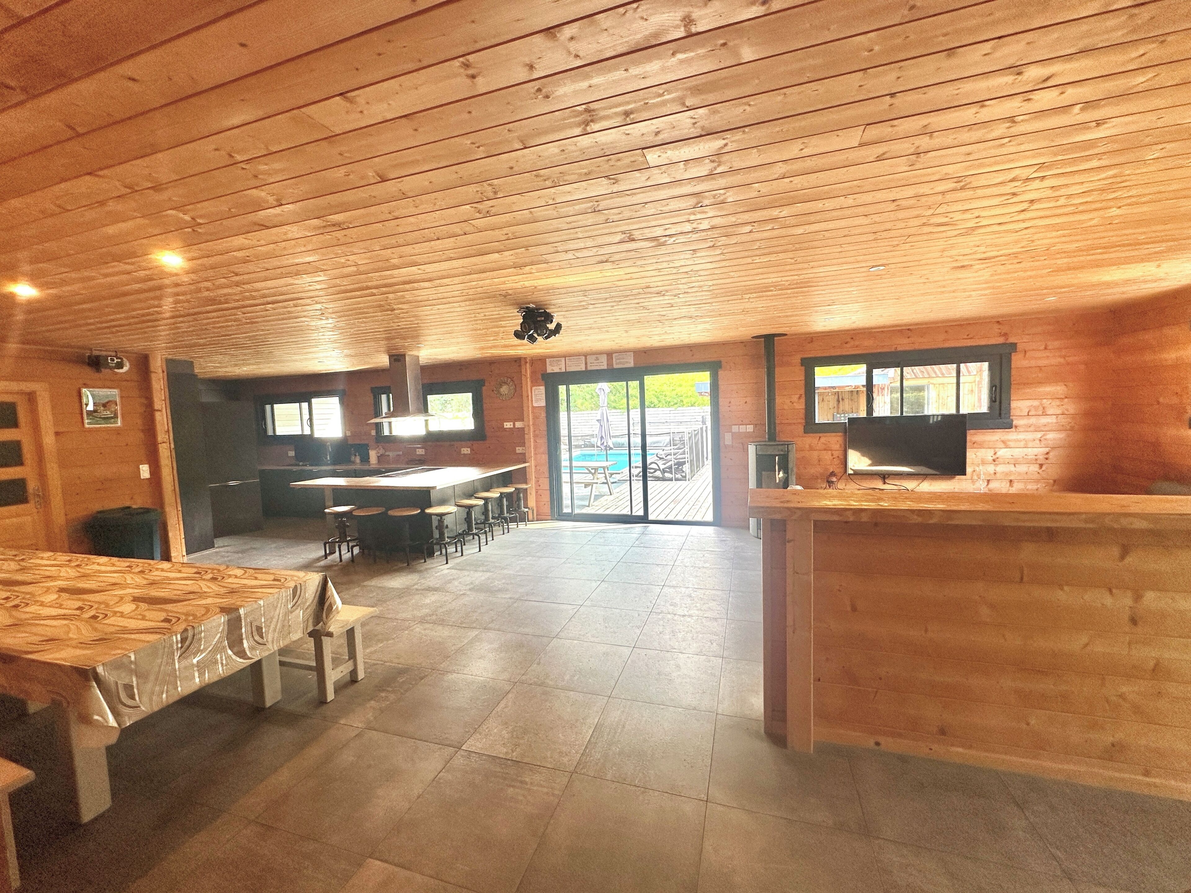Amplo espaço interno com teto de madeira, área de bar com banquetas, cozinha com uma ilha grande e vista para um deck externo por uma porta de vidro.