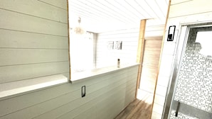 A narrow hallway with light-colored walls and wooden flooring. There is a window on the left side, a door on the right, and a small shelf with a bottle.
