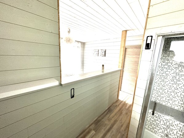 Un couloir étroit aux murs de couleur claire et au plancher en bois. Il y a une fenêtre sur le côté gauche, une porte sur le côté droit et une petite étagère sur laquelle se trouve une bouteille.