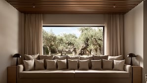 Un salon moderne avec un canapé beige, deux lampadaires noirs et une grande fenêtre donnant sur un paysage d’oliviers.