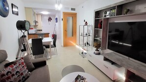 A modern living room with a large flat-screen TV, a dining area with a table and chairs, and a kitchen in the background. - Marvel Apartment with Pool near Playa Vera (Vera Playa)