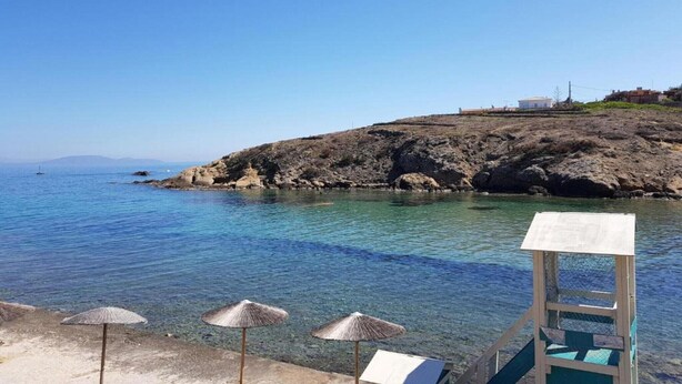 A clear blue sea with rocky terrain, a lifeguard tower, and beach umbrellas. - Apartment in Rafina with Garden & BBQ (Rafina)
