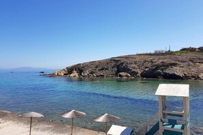 A clear blue sea with rocky terrain, a lifeguard tower, and beach umbrellas. - Apartment in Rafina with Garden & BBQ (Rafina)