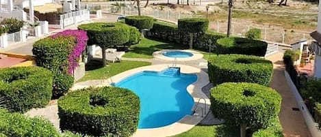 A pool area with two swimming pools, surrounded by well-manicured hedges and a white building with balconies.