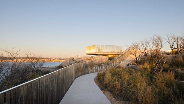 Een modern gebouw met een houten looppad er naartoe, omgeven door natuur en een heldere lucht.