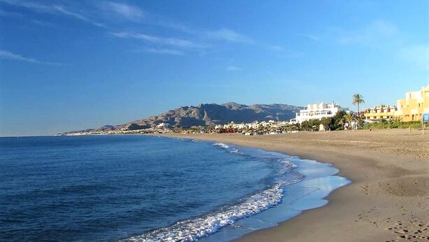 A beach with clear blue water, a sandy shore, and buildings in the background.
