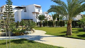 A modern residential building with a flat roof, multiple balconies, and a well-maintained garden with palm trees and shrubs.