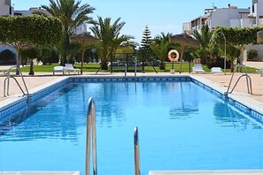 A rectangular swimming pool with a metal ladder in the foreground, surrounded by palm trees and lounge chairs.