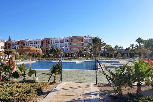 A resort with a swimming pool, palm trees, and a row of multi-story buildings with balconies.