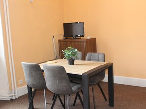Uma sala de jantar com mesa, cadeiras, um vaso de planta, televisão sobre um móvel e um radiador branco.