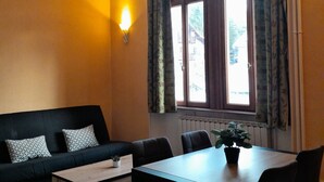 A room with a black sofa, a wooden table, and a window with curtains. - T2 Apartment in Castle, 4 people, pool, pets allowed (Jausiers)
