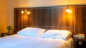 Et hotelværelse med en træsenggavl, to sengeborde med lamper, en seng med hvide sengetøj, et lille sengetablet med en plante, og et natbord med bøger.