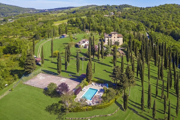 An aerial view of a property with a swimming pool, tennis court, and a large building surrounded by trees.