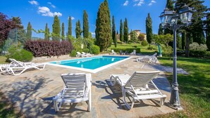 A swimming pool with lounge chairs, a lamp post, and tall trees in the background. - Villa La Chiesina (Arcidosso)