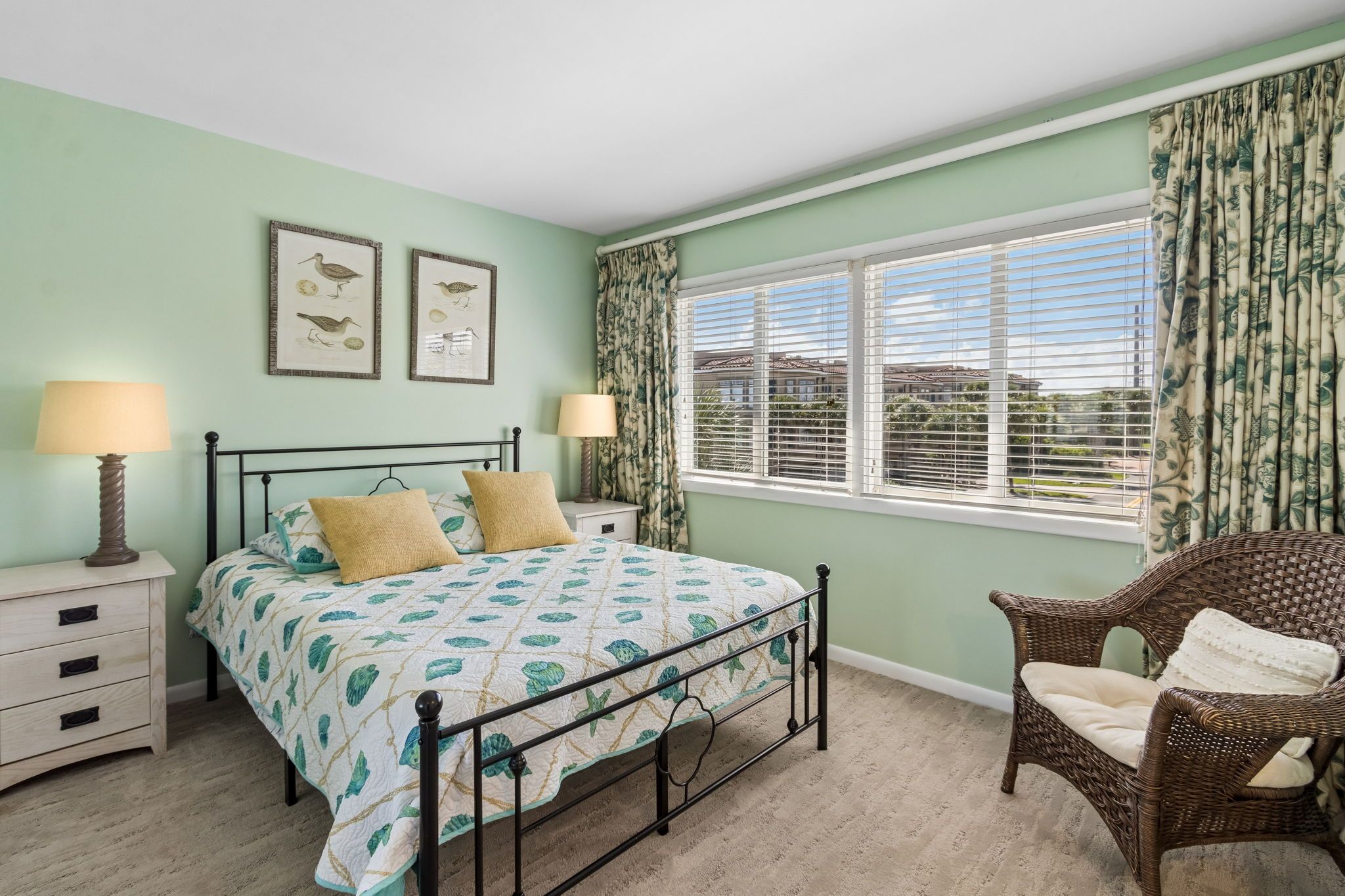 A bedroom with a bed, two bedside tables with lamps, a wicker chair, a window with blinds, and two framed pictures on the wall.