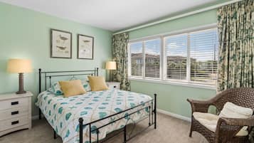 A bedroom with a bed, two bedside tables with lamps, a wicker chair, a window with blinds, and two framed pictures on the wall.