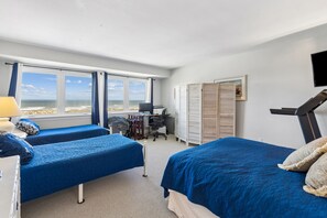 A hotel room with two beds, a desk, a chair, a TV, and a view of the beach.