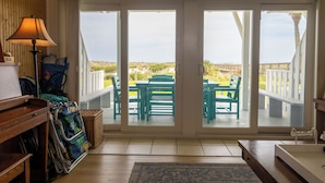 A room with a wooden desk, a lamp, a wicker basket, and a view of an outdoor patio with green chairs and a table. - Lovely Beach house in Amelia for 8 (Fernandina Beach)