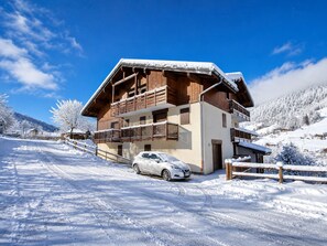 Eine zweistöckige Holzhütte mit Balkonen, ein Auto davor und ein verschneiter Berg im Hintergrund.