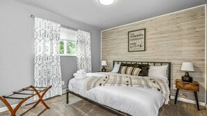 A bedroom with a bed, two bedside tables with lamps, a window with patterned curtains, and a wooden wall panel. - Modern Family Haven • Fenced Yrd • Grill/Firepit (Westminister)