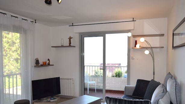 Una sala de estar moderna con un sofá, una mesa de centro y una televisión. Hay estanterías con objetos decorativos y un amplio ventanal con vistas al exterior.