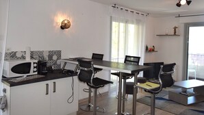 Una cocina moderna con armarios blancos, encimera negra y microondas. Hay cuatro taburetes de bar y una mesa de comedor con sillas. Se ve una lámpara de pared decorativa y una ventana con cortinas.