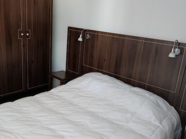 Quarto com cabeceira de madeira, colcha branca e mesa de cabeceira com abajur.