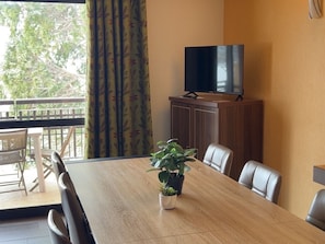 Une salle de conférence avec une table en bois, des fauteuils en cuir, une plante en pot, un téléviseur posé sur un meuble et une vue sur les arbres à travers une fenêtre ornée de rideaux.