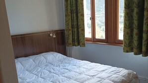 A single-bed room with a wooden headboard, a window with green patterned curtains, and a white bedspread. - T3 apartment in castle with pool and WIFI, pets allowed, 6 people (Jausiers)